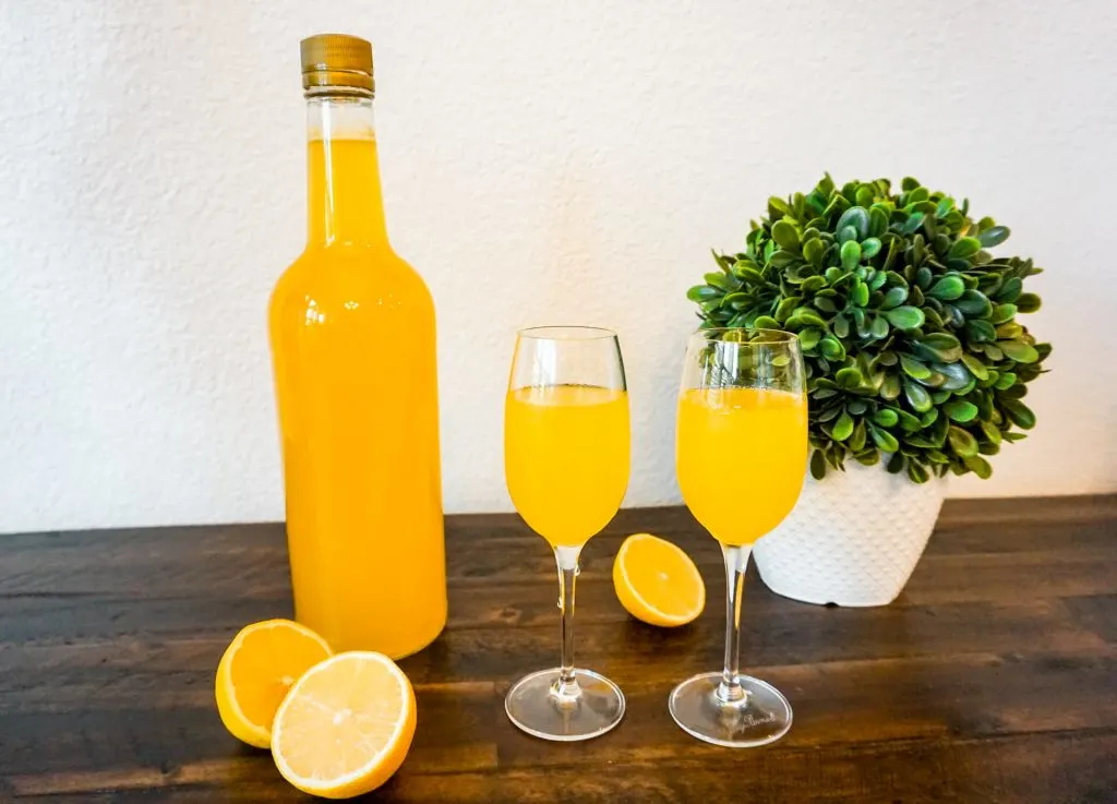 A bottle of homemade limoncello next to two glasses full of limoncello and sliced lemons around it.
