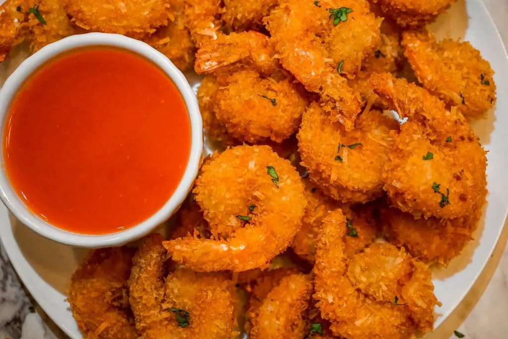 Coconut crusted shrimp with parsley on top and surrounded by a side of sweet red bell pepper chili sauce.