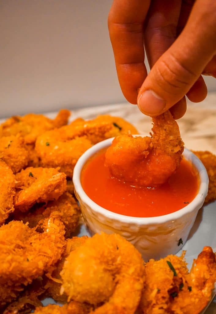 A hand dipping coconut shrimp into a ramekin full of a sweet red bell pepper chili sauce.