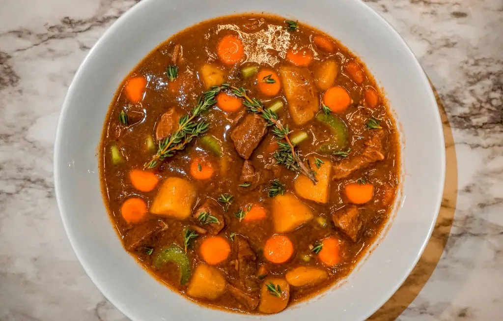 A bowl of Instant Pot Guinness Beef Stew - sprigs of fresh thyme, cubes potatoes, carrots, celery, and chuck roast in a beef broth.