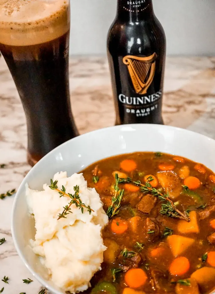 A glass and bottle of Guinness behind a bowl of Instant Pot Guinness Beef Stew with mashed potatoes.