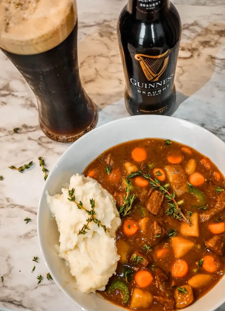 A glass of Guinness alongside a bottle of Guinness and a bowl of Instant Pot Guinness Beef Stew with mashed potatoes.