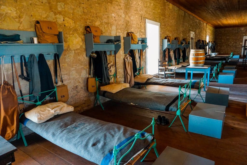 A restored building from Fort Concho's soldier's quarters in San Angelo. There are beds lined in a row with chests and other soldiers' belongings.