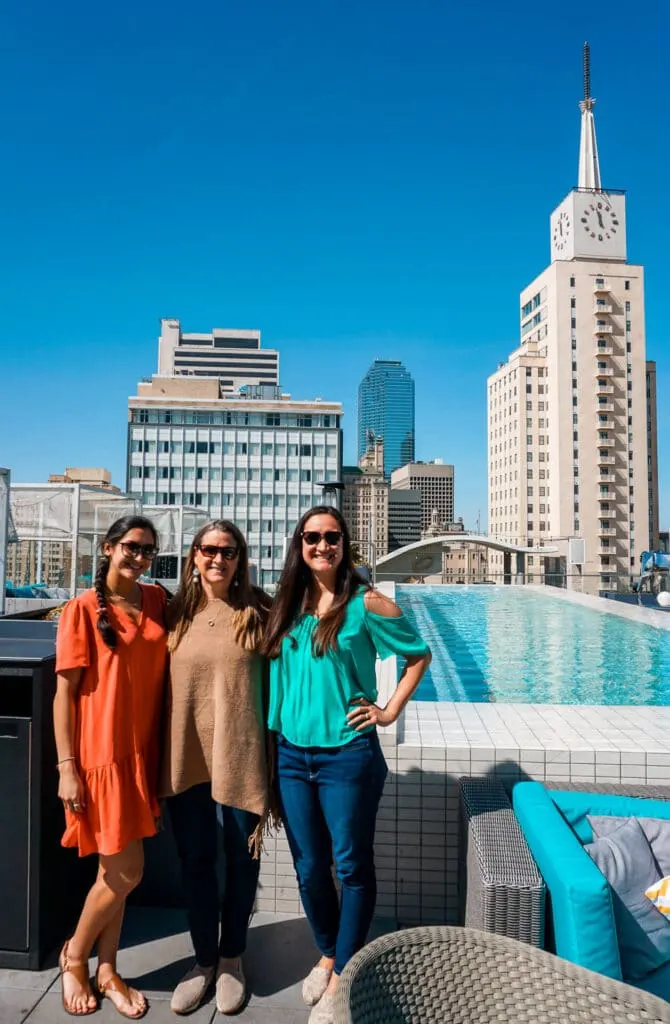 Three women enjoying a girl's weekend in Dallas at the Statler Hotel's rooftop pool with skyscrapers in the background.