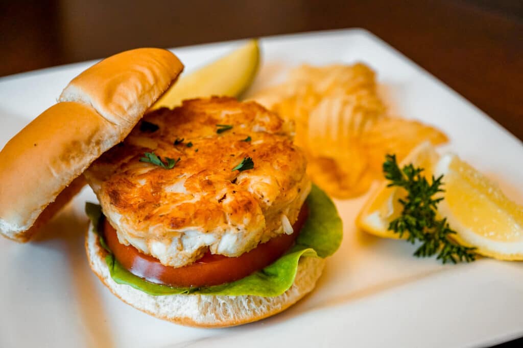 A crab cake sandwich on a plate with lemon wedges, a pickle, and kettle chips.