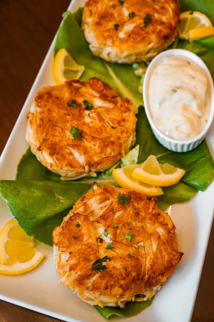 A platter of three Maryland Crab Cakes on a bed of lettuce with lemon triangles and a side of tartar sauce.