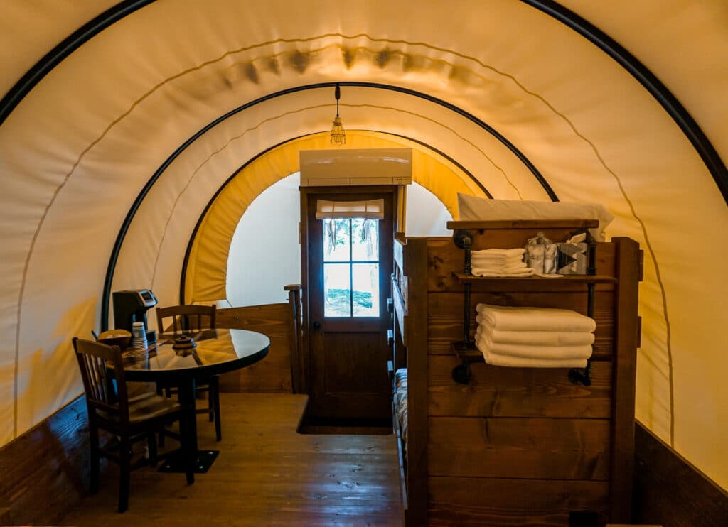 View of inside a Conestoga Covered Wagon from the king-sized bed. Pictured is a nook filled with towels from a bunk bed and a wagon wheel table.
