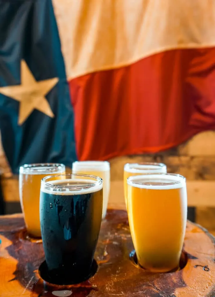 A beer flight of four glasses on a wooden platter with the Texas flag in the background from The Q & Brew.