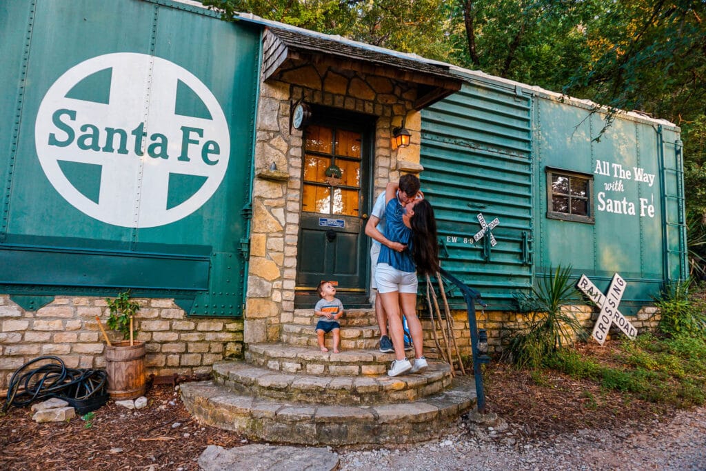 A couple kissing on the steps while a baby looks up at them in front of a green vintage Santa Fe Railcar at Country Woods Inn in Glen Rose, Texas.