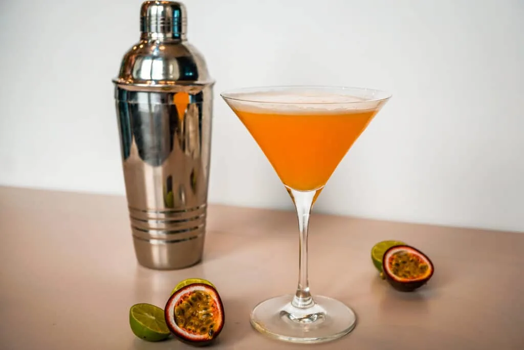 A cocktail shaker, Passion Fruit Pisco Sour cocktail in a martini glass and sliced passion fruit and limes.