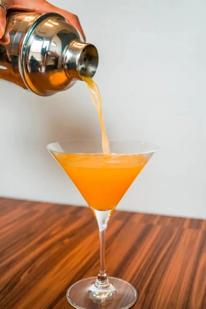 Someone pouring an orange Passion Fruit Pisco Sour from a cocktail shaker and into a martini glass.