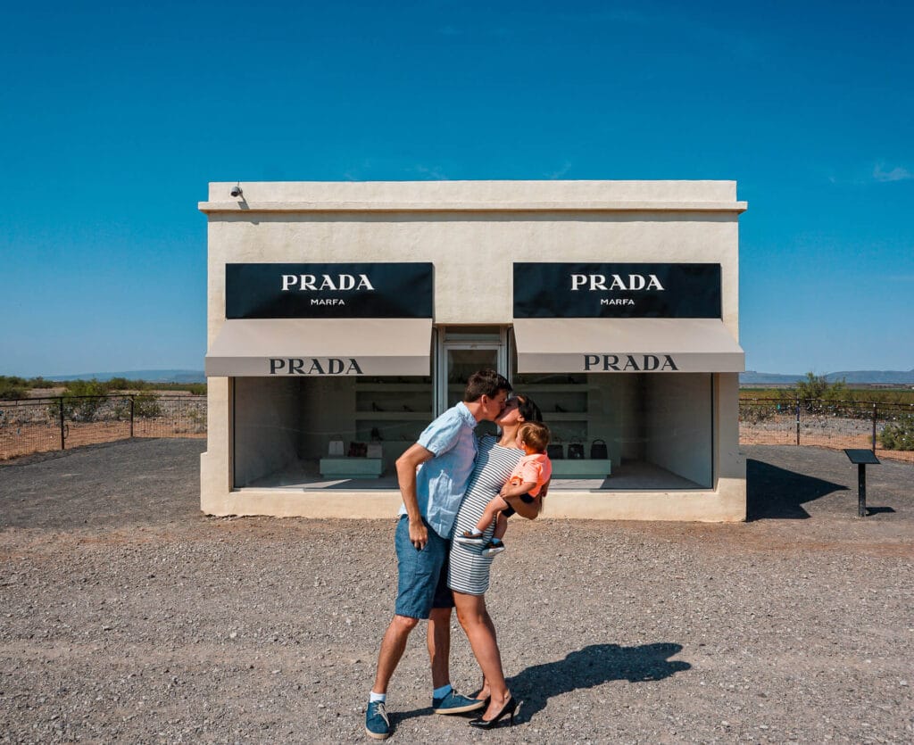 A couple holding their baby son while kissing in front of Prada Marfa (a permanent art installation) - one of the most iconic roadside attractions and best things to do in West Texas. 