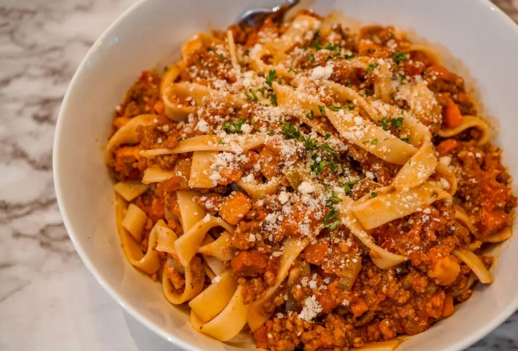 A bowl of Bolognese Sauce tossed with Tagliatelle pasta and a sprinkle of grated cheese and parsley on top.