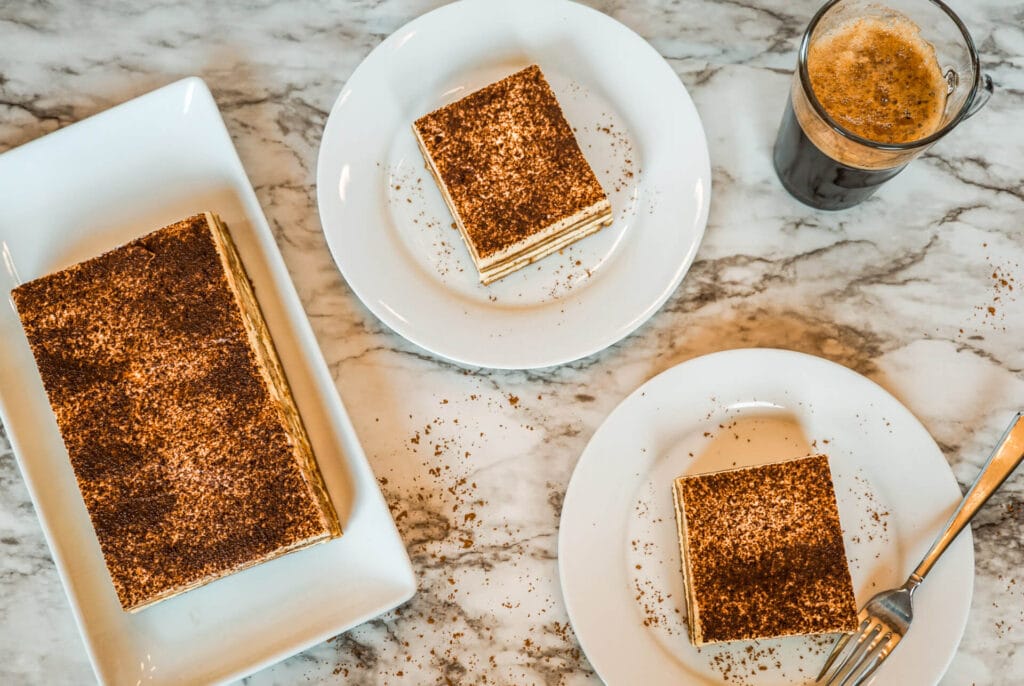 Birdseye view of a large Tiramisu cake and two small plates with slices of Tiramisu and a glass of coffee on a marble table.