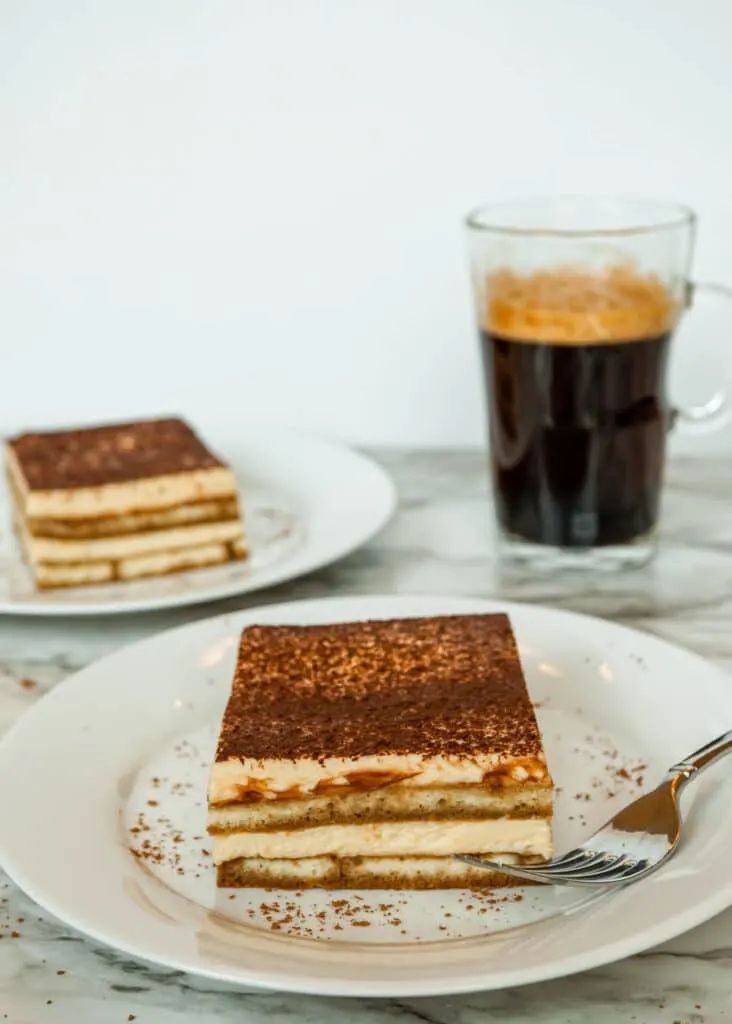 A slice of safe to eat when pregnant Tiramisu on a plate with a fork next to it and a glass of coffee in the background.