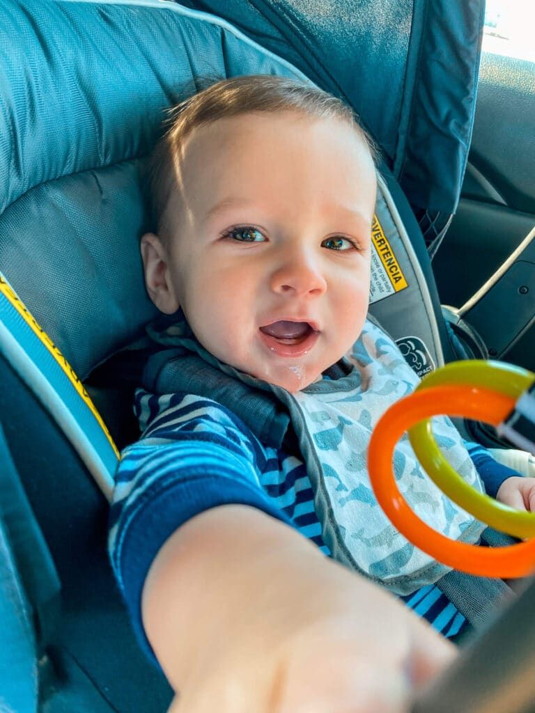 Road tripping with a baby at 9 months old - baby boy looking and reaching out at camera while seated in a car seat.