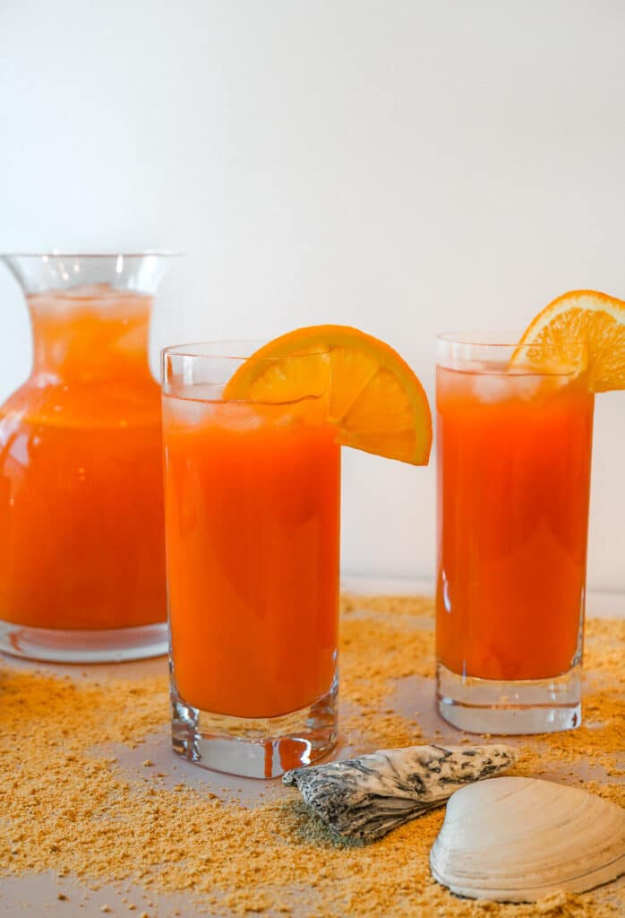 A pitcher and two glasses filled with a reddish orange rum punch cocktail - the Rum Swizzle. 