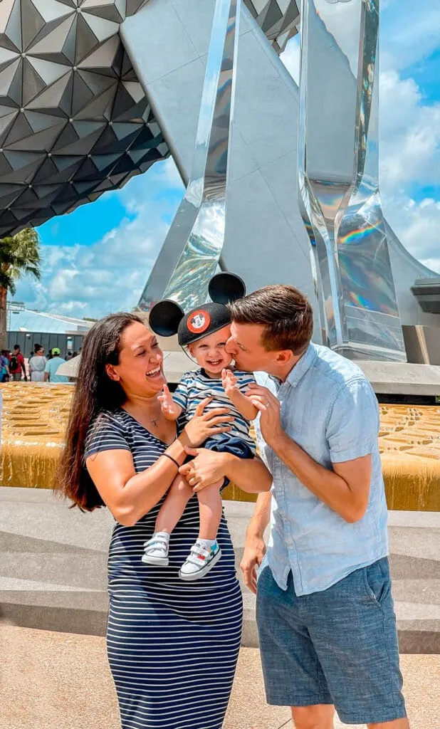 A mom and dad at Epcot with a baby (kissing their son) in front of geosphere.