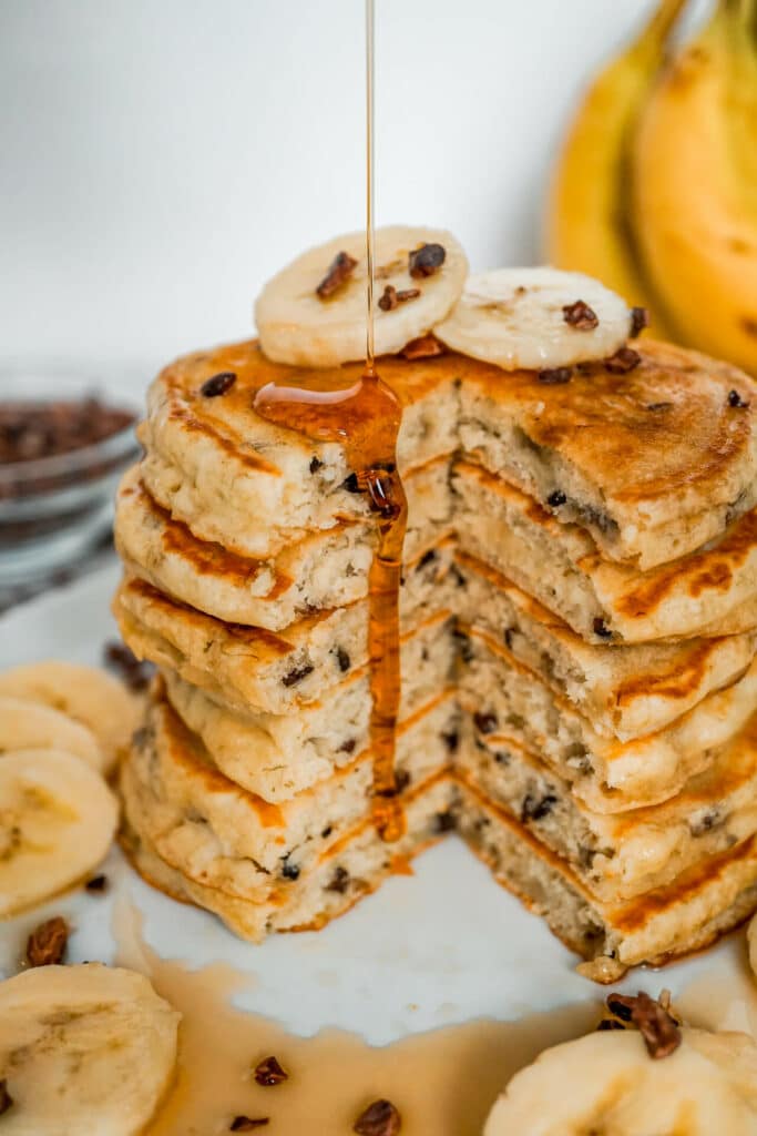 A stack of banana cacao nib pancakes with a slice removed and maple syrup dripping down the side.