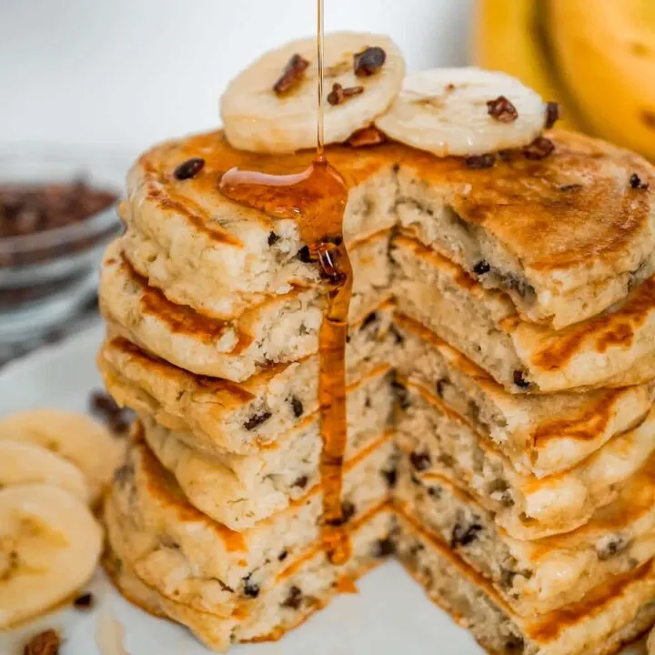 A stack of banana cacao nib pancakes with a slice removed and maple syrup dripping down the side.