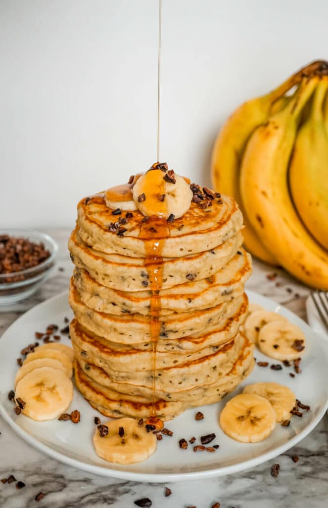 Maple syrup being poured over a stack of healthy, fluffy banana cacao nib pancakes.