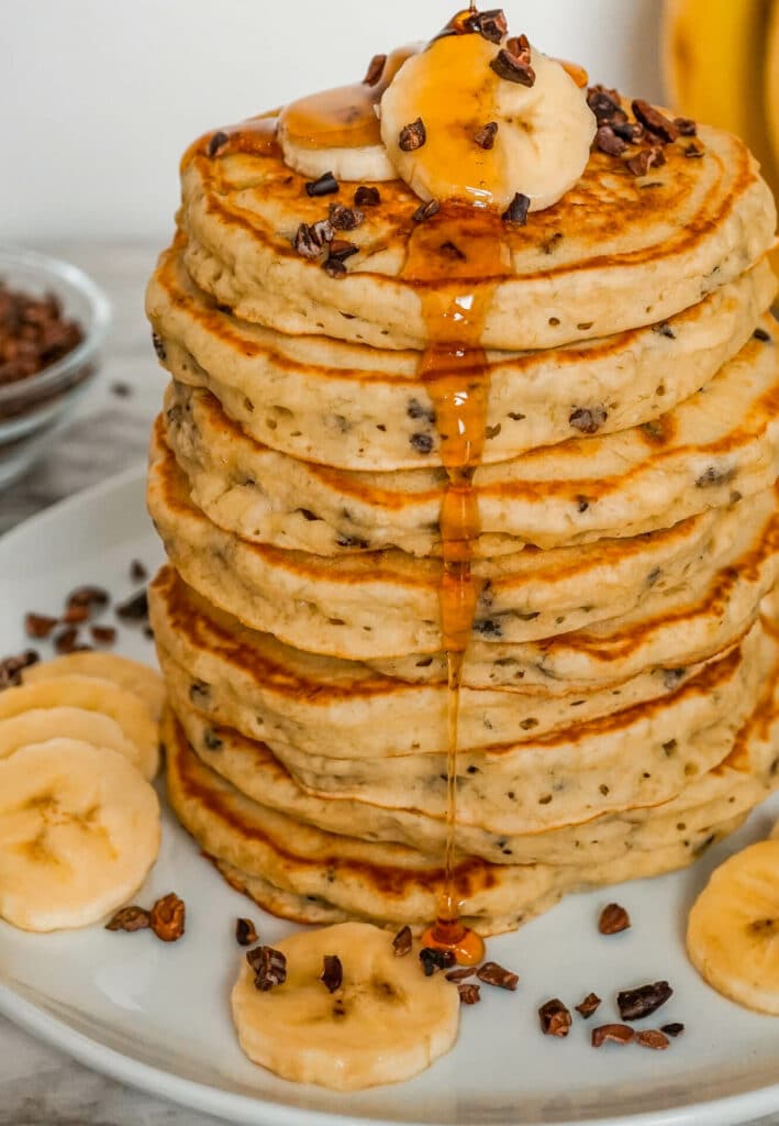 A stack of banana cacao nib pancakes with maple syrup dripping down.