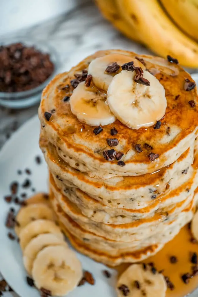 A stack of banana cacao nib pancakes with sliced bananas on top and around the plate.