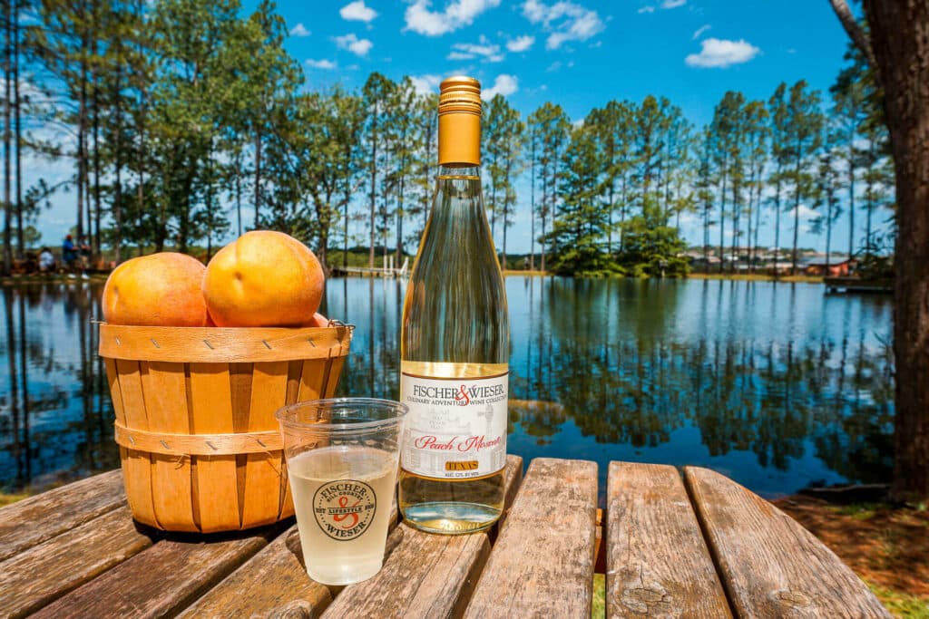 A bucket of peaches, a glass of white wine, and a bottle of wine from Das Peach Haus with a pond in the background.