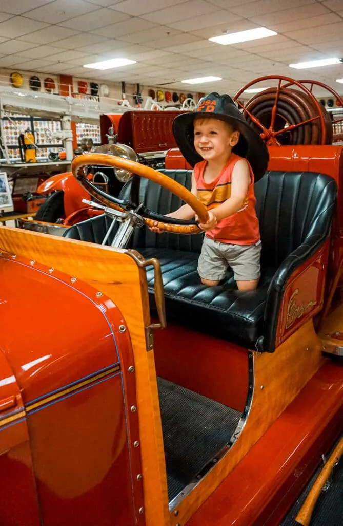 A toddler pretending to drive a restored firetrucks with a black firefighter hat at the Oklahoma Firefighters Museum.