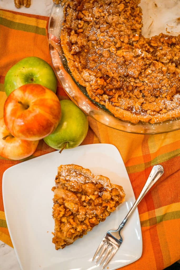Birdseye view of a slice of Caramel Apple Walnut Pie with a Streusel Topping beside a pie dish of more apple pie and a stack of apples.