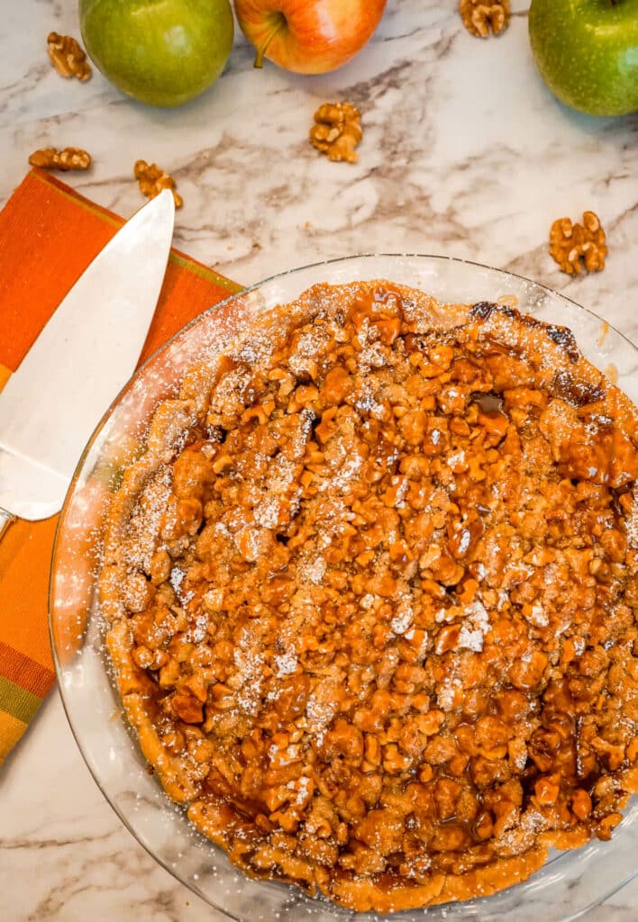 A Caramel Apple Walnut Pie with a Streusel Topping with walnuts around it and a pie serving utensil. 
