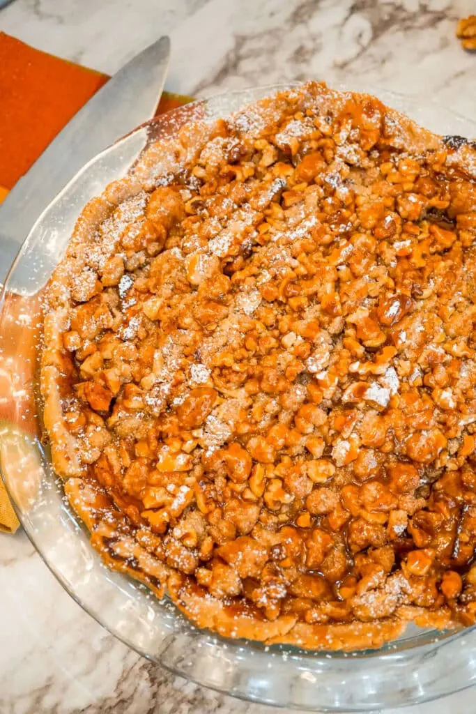 A whole Caramel Apple Walnut Pie with a Streusel Topping in a glass pie dish with a pie serving utensil next to it.