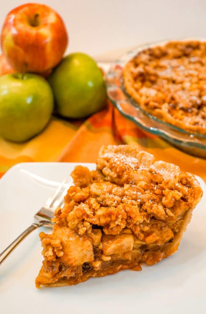 A slice of Caramel Apple Walnut Pie with a Streusel Topping on a white plate with a fork beside it and a stack of apples in the background.