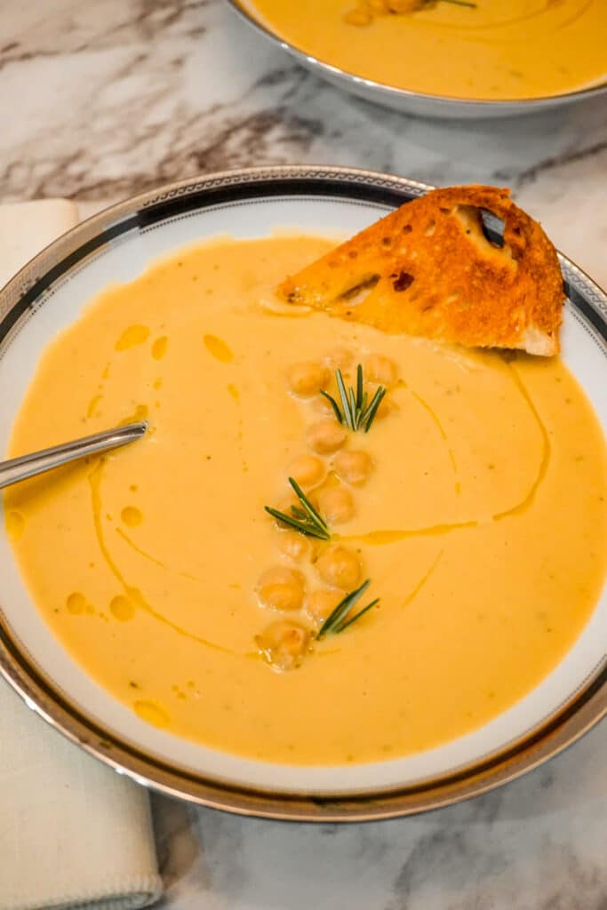A bowl of Creamy Tuscan Chickpea Soup with olive oil drizzled over it and a slice of bread, sprigs of rosemary, and chickpeas on top. 
