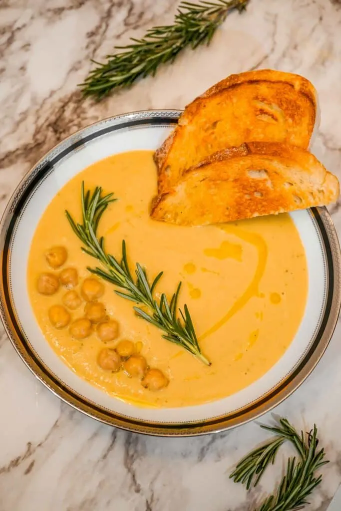 A bowl of Creamy Tuscan Chickpea Soup with olive oil drizzled over it and a slices of bread, sprig of rosemary, and chickpeas on top. 