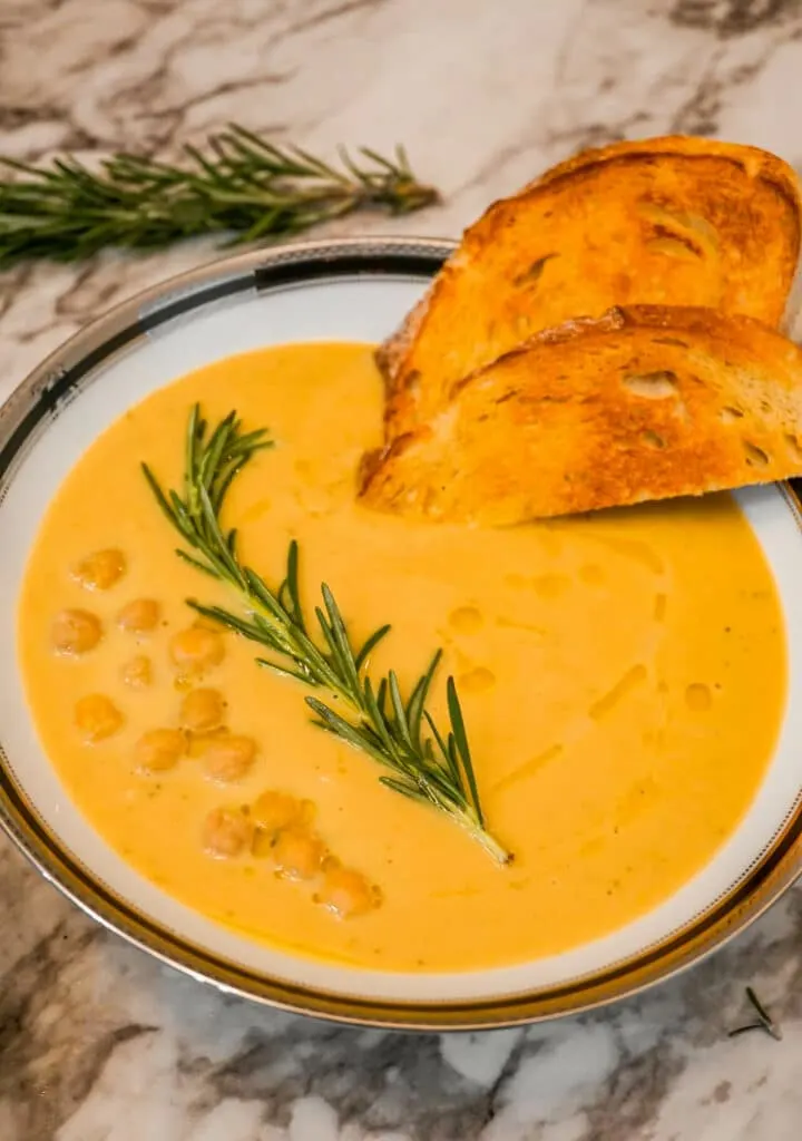 A bowl of Creamy Tuscan Chickpea Soup with olive oil drizzled over it and a slices of toasted bread, a rosemary sprig, and chickpeas on top. 