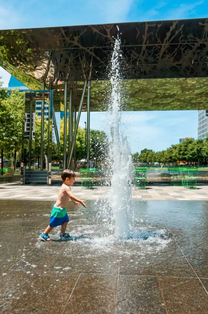 A toddler in swim trunks running through a water fountain at Kyle Warren Park - one of the best things to do in Dallas with toddlers.