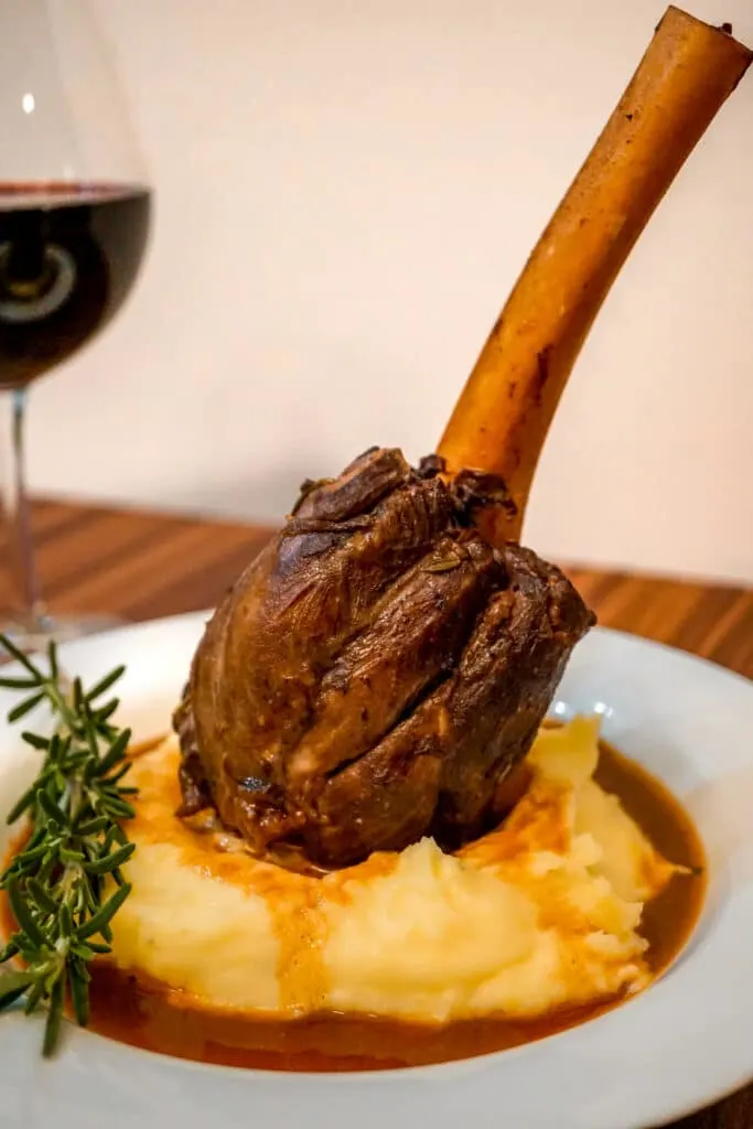 A Braised Lamb Shank with exposed bone standing on a bed of creamy mashed potatoes and a sprig of rosemary.