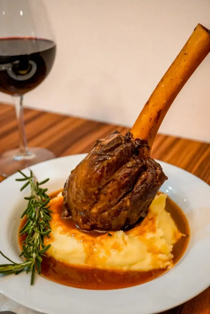 Instant Pot Braised Lamb Shanks standing up right with the meat part down on a bed of mashed potatoes and the exposed bone up. The Red Wine Jus is around the mashed potatoes with a sprig of rosemary and glass of red wine in the back.