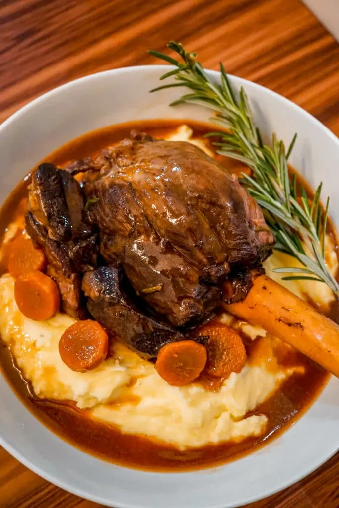 A glossy look on Instant Pot Braised Lamb Shanks with Red Wine Jus, sliced carrots, rosemary, and mashed potatoes.