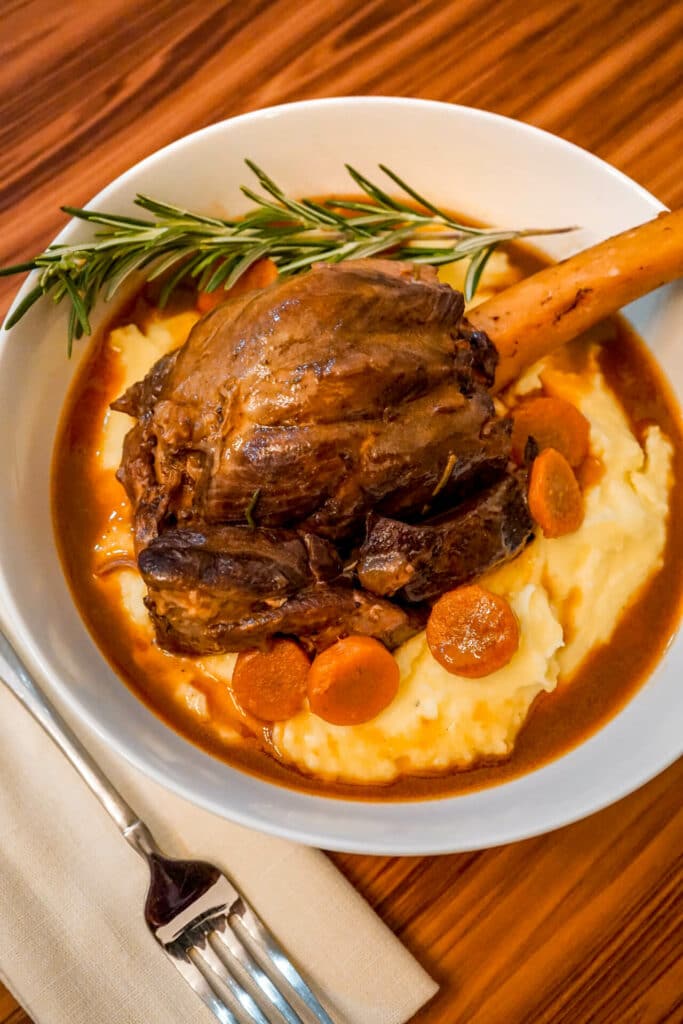 Instant Pot Braised Lamb Shanks with a Red wine Jus on a bed of creamy mashed potatoes, sliced carrots, and a sprig of rosemary.