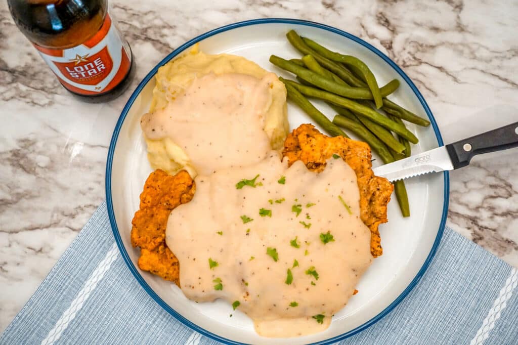 A plate of Texas Chicken Fried Steak and mashed potatoes smothered in a Beer Gravy with chopped parsley,  and green beans.