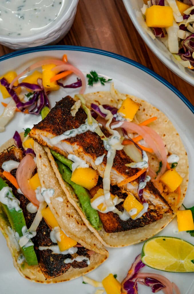 Two strips of Blackened Fish Tacos with Mango Slaw, pickled red onions, sliced avocado, and drizzled lime crema. 