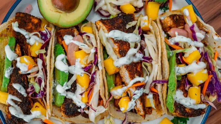 Four Blackened Fish Tacos with Mango Slaw, pickled red onions, sliced avocado, and drizzled lime crema.