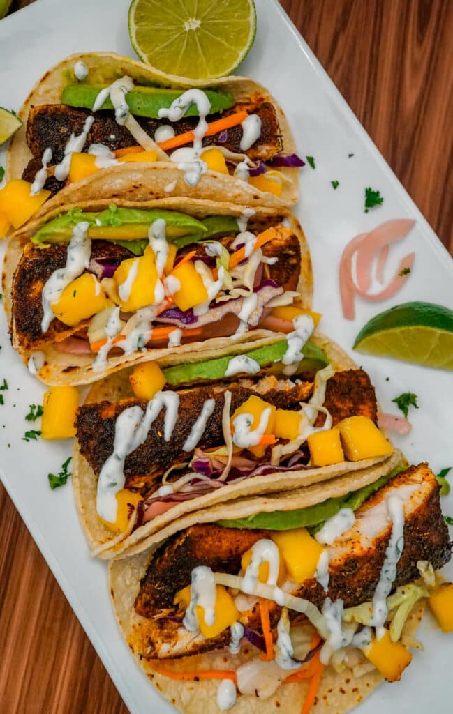 A road of blackened fish tacos with chunks of mango, pickled red onion, sliced avocado, and lime crema drizzled on top.