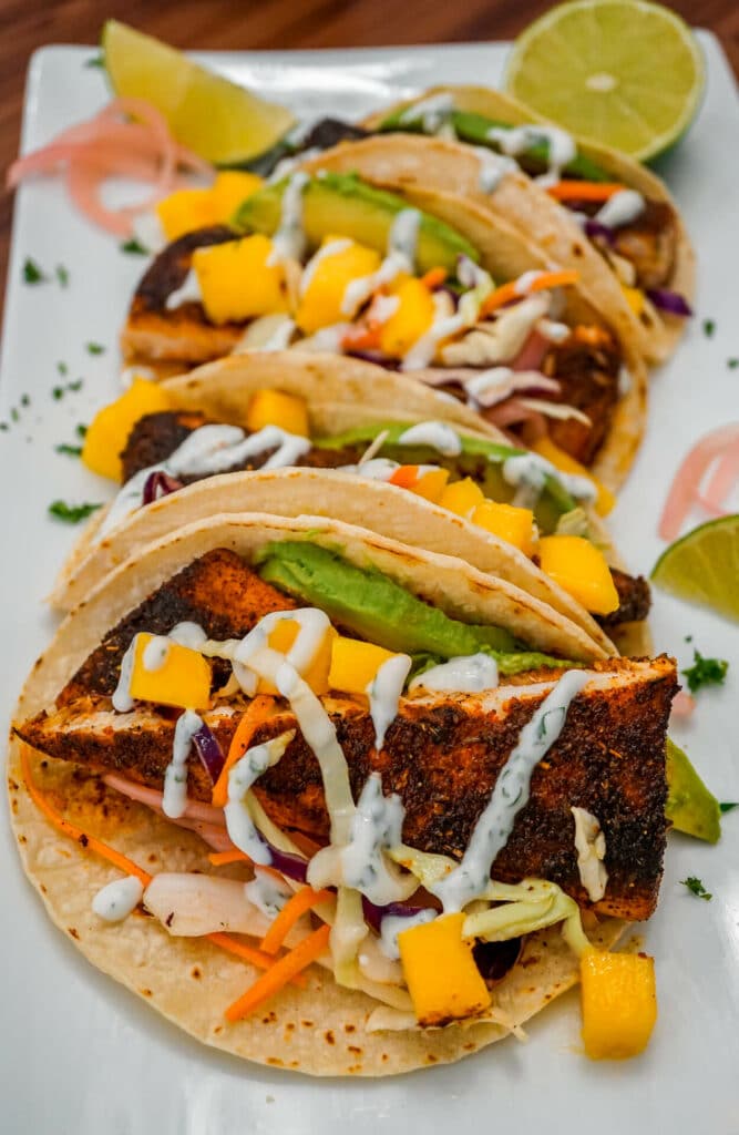A row of Blackened Fish Tacos with chunks of mango, cabbage slaw, pickled red onions, sliced avocado, and drizzled lime crema. 