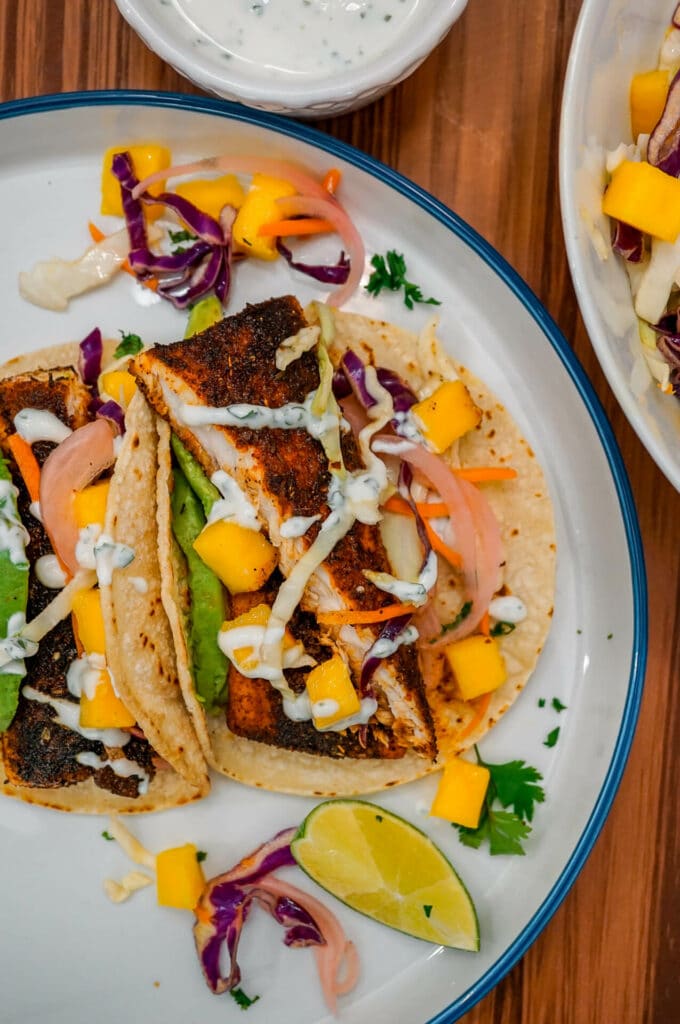 A plate of Blackened Fish Tacos with Mango Slaw, pickled red onions, sliced avocado, and drizzled lime crema. 