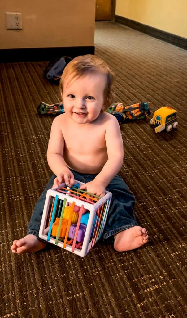 A baby boy sitting in a hotel room shirtless playing with a cube and textured blocks - one of the best travel toys for babies.