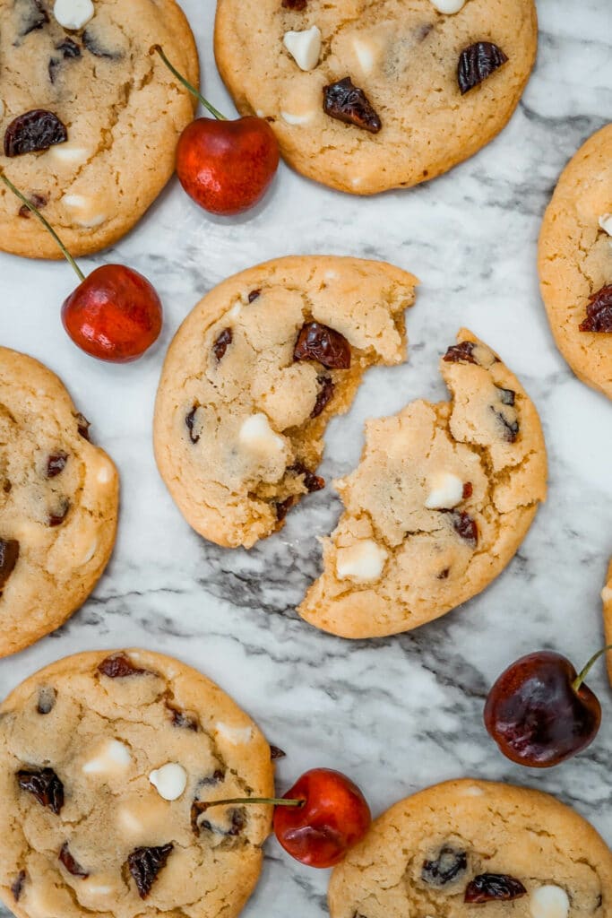 One White Chocolate Cherry Cookies in two pieces with other cookies and fresh cherries surrounding it.