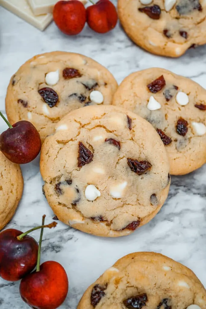 Three White Chocolate Cherry Cookies on top of each other with other cookies and fresh cherries surrounding them.